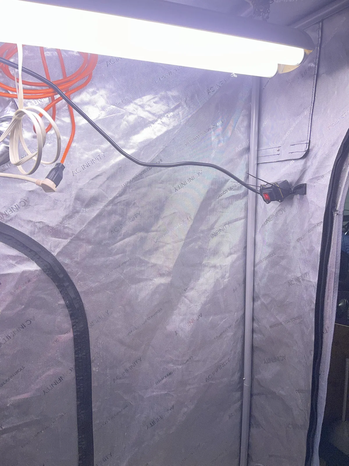 The old non-UV fish tank light hanging from chains, its black cord running along the tent wall to the external red rocker switch zip-tied to the frame near the door, and coiled extension cords hung on the wall.