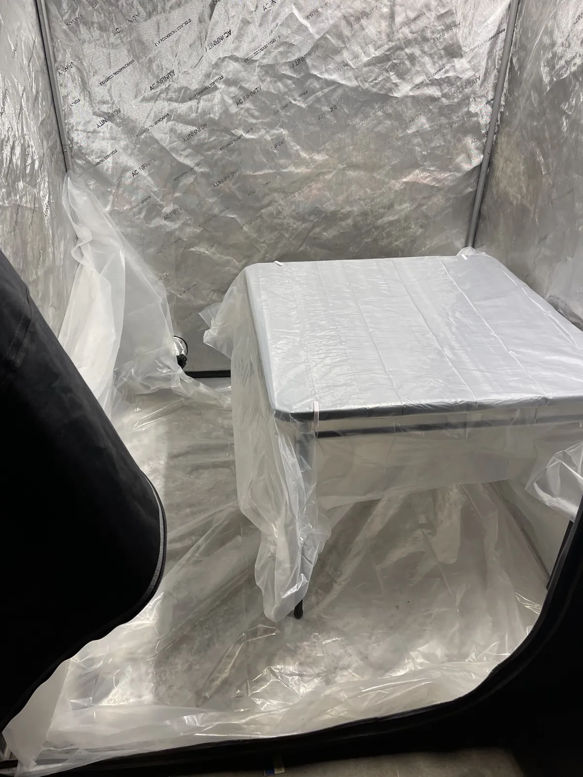 The tent with plastic over the folding table and across the floor, and white clips holding the plastic at the table's edge.