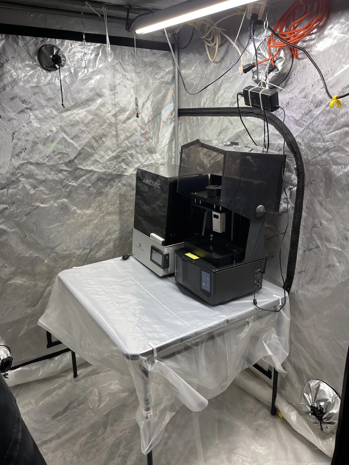 An angled view of the same table. White clips hold the plastic at the table's edge and a leg, a power adapter is zip-tied to the tent wall above the machines, and the Saturn's hood is open.