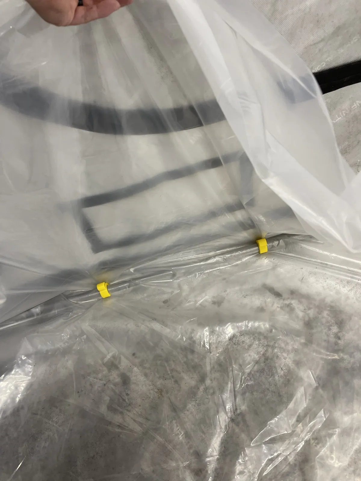 Lifting the liner away from the wall to show two yellow clips holding the plastic to the lower frame tube.