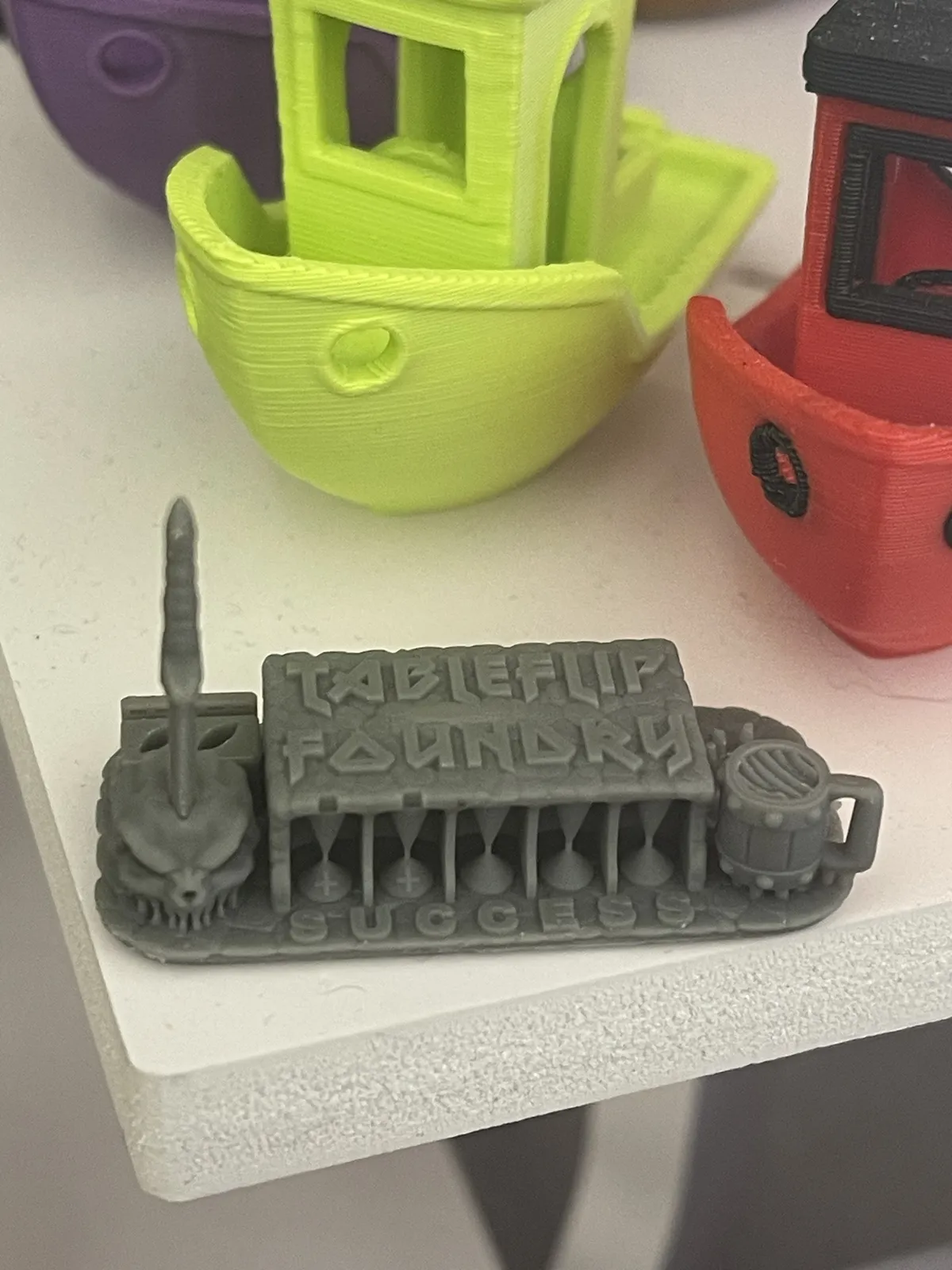 The Cones of Calibration v3 in SUNLU Dark Grey resin, top-down: the TableFlip Foundry roof, the skull and spike at left, the mug at right, and SUCCESS along the front of the base.