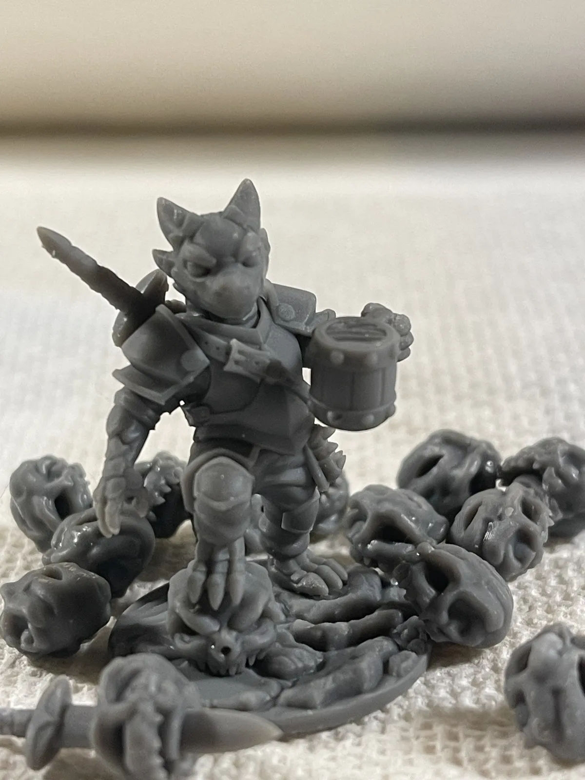 Puck in Dark Grey resin, front view: armored, holding a banded tankard, with a sword over his shoulder, standing on a skull-strewn base. The loose skulls and the sword in front came off other calibration prints.